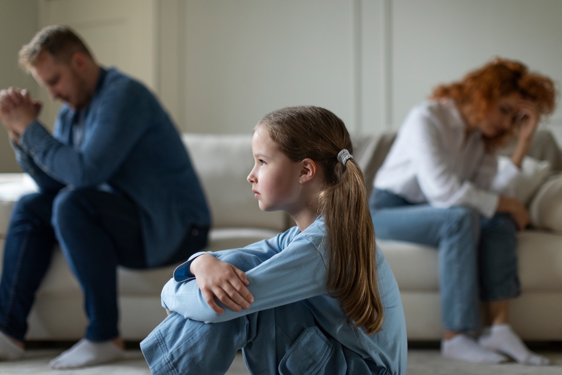 A young girl sits apart while two adults sit distressed in the background.